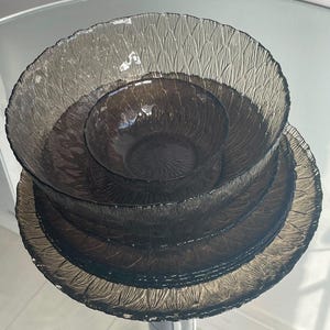 Peut inclure: Un ensemble de trois bols en verre gris foncé emboîtables présentant un motif texturé en forme de feuilles sur l'extérieur. Les bols sont empilés verticalement, avec le plus petit en haut, suivi d'un bol moyen et d'un grand bol à la base.
