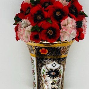 May include: A decorative ceramic wall pocket vase featuring a flared rim with gold trim. The body displays intricate black, white, and gold patterns with floral motifs. It is filled with a dense arrangement of vibrant red and white artificial flowers.