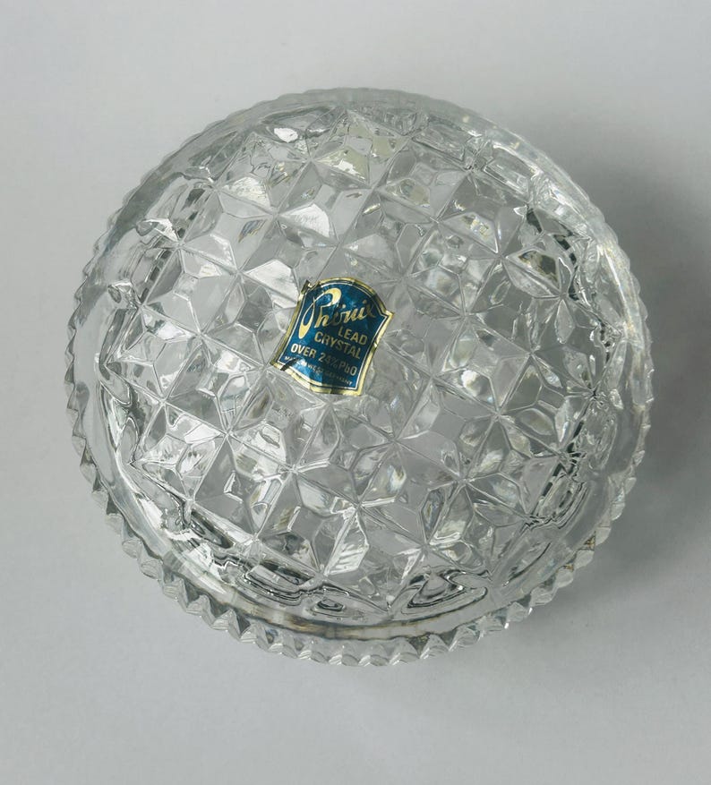 May include: Clear lead crystal bowl with a diamond pattern. The bowl has a scalloped edge and a blue and gold label that reads "Lead Crystal Over 24% Pbo".
