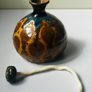 Puede incluir: Pequeña lámpara de aceite de cerámica redonda, con un patrón moteado marrón y amarillo. La parte superior es marrón oscuro con un esmalte azul. Una mecha de algodón blanco está unida a una pequeña cuenta oscura.