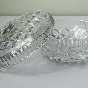 May include: A clear, cut-crystal trinket box with a lid, featuring a diamond pattern and a ribbed edge. The lid is partially open, revealing the interior of the bowl. A small blue and gold label is visible on the lid.