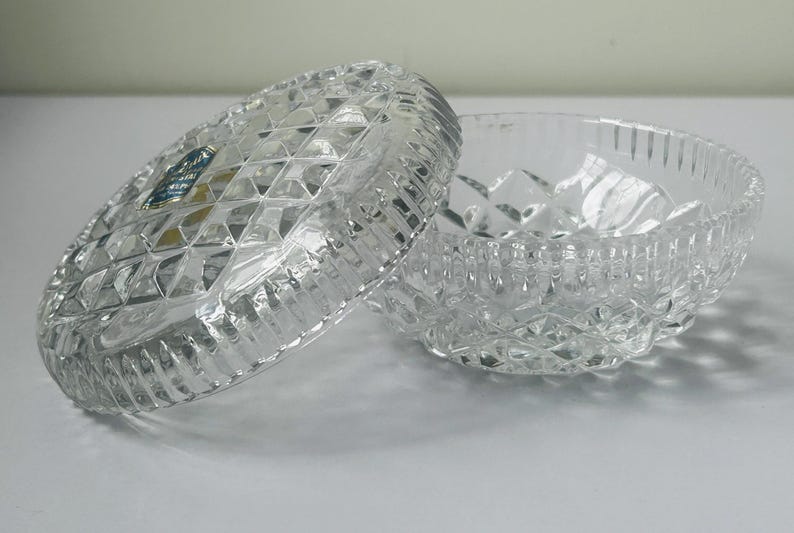 May include: A clear, cut-crystal trinket box with a lid, featuring a diamond pattern and a ribbed edge. The lid is partially open, revealing the interior of the bowl. A small blue and gold label is visible on the lid.