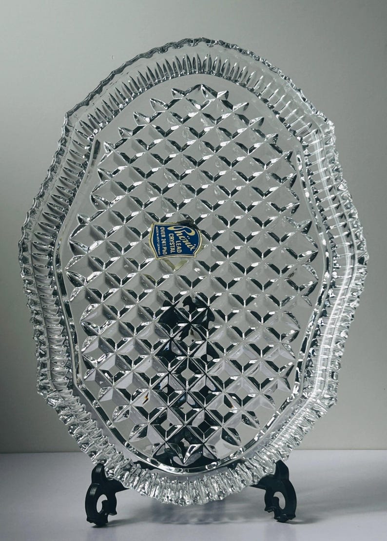May include: Clear lead crystal serving tray with a diamond pattern and scalloped edges. The tray has a blue and white label that reads "Phoenix Crystal Over 24% Pbo". The tray is displayed on a black metal stand.