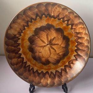 Pode incluir: Um prato redondo de cerâmica com um padrão floral central de pinceladas castanho-escuras, rodeado por um anel laranja-acastanhado mais claro e uma borda exterior de pinceladas festonadas castanho-escuras sobre um fundo bege, num suporte preto.