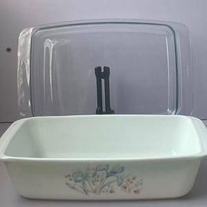 May include: A rectangular white baking dish with a floral pattern featuring blue irises and small orange accents on the front. The dish includes a clear glass lid with integrated handles, positioned upright behind the main white ceramic vessel.