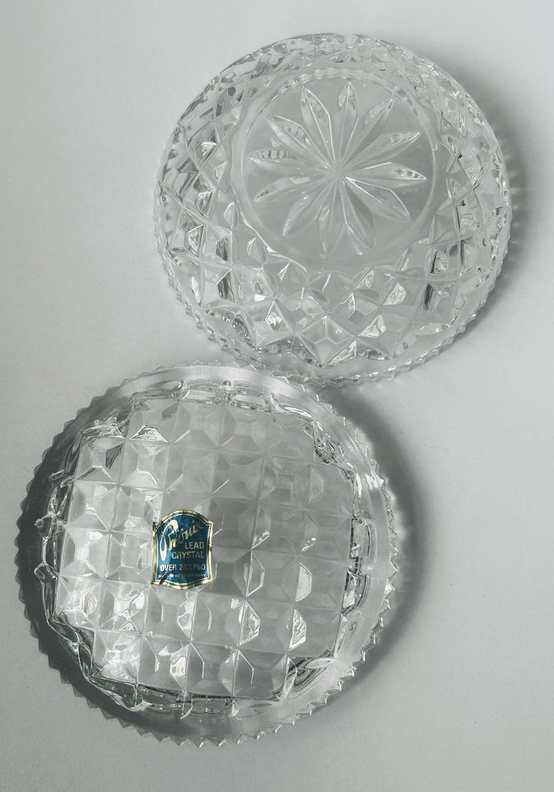 May include: Two clear, cut-glass bowls with diamond and starburst patterns. One bowl has a label that reads "Lead Crystal Over 24% Pbo". The bowls are round with a scalloped edge, and are likely decorative or serving pieces.