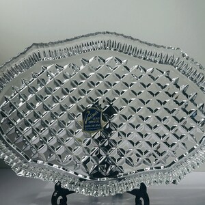 May include: Clear lead crystal serving tray with a diamond pattern and a scalloped edge. The tray has a label that reads "Phoenix Lead Crystal Over 24% Pbo". The tray is oval-shaped and sits on a black metal stand.
