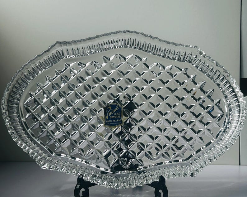 May include: Clear lead crystal serving tray with a diamond pattern and a scalloped edge. The tray has a label that reads "Phoenix Lead Crystal Over 24% Pbo". The tray is oval-shaped and sits on a black metal stand.