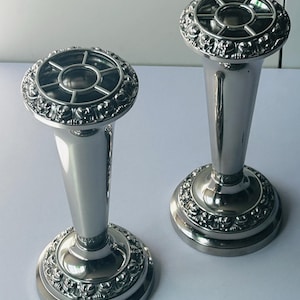 May include: A pair of silver-toned metal flower frogs or bud vases with a tapered cylindrical body. Each piece features a circular base and a top rim decorated with a raised floral pattern, topped with a removable metal grid insert for stems.
