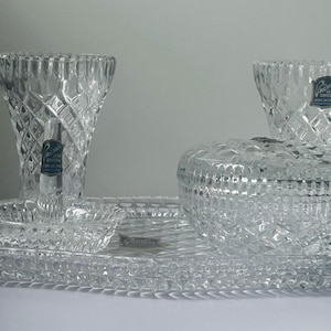 May include: A clear crystal vanity set with diamond-cut patterns. The set includes a vase, two small bowls, a lidded jar, and an oval tray. Each piece has a label that reads "Princess House".
