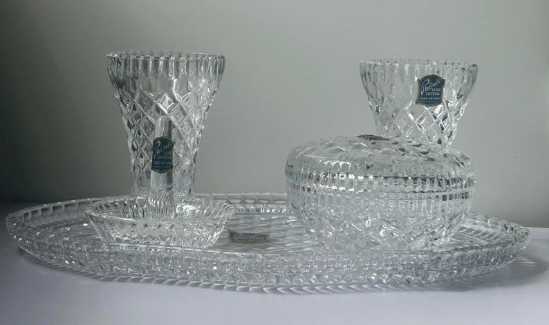 May include: A clear crystal vanity set with diamond-cut patterns. The set includes a vase, two small bowls, a lidded jar, and an oval tray. Each piece has a label that reads "Princess House".