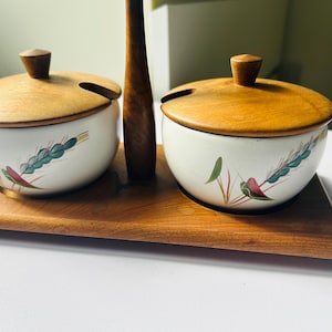 May include: A wooden serving set with two lidded ceramic bowls, each with a wheat design, on a wooden tray. The lids and tray are brown, the bowls are white with green, pink, and blue accents. A charming kitchen accessory.