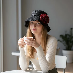 May include: A black sequined cloche hat with a large red rose on the side. The hat is worn by a person with long blonde hair, holding a white cup. The person is wearing a cream-colored sweater.