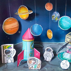 Puede incluir: Diorama de papel colorido del sistema solar, con un cohete, astronautas, planetas y un perro con traje espacial. Los planetas están suspendidos desde arriba, sobre un fondo azul oscuro.