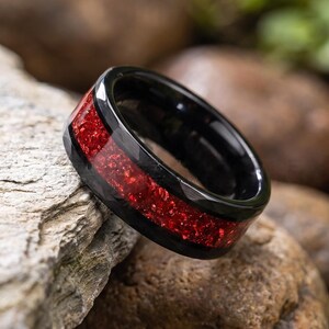 Könnte beinhalten: Ein schwarzer Ring mit einem breiten, vertieften Mittelband, das mit einem leuchtenden, schimmernden roten Material gefüllt ist. Der Ring hat eine glatte, polierte Oberfläche an den Kanten, was einen deutlichen Kontrast zwischen dem dunklen Metall und der roten Einlage erzeugt.