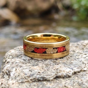 Puede incluir: Un anillo de banda en tono dorado con una incrustación central de fragmentos rojos y dorados metálicos sobre un fondo oscuro. El anillo tiene un acabado pulido con bordes lisos y elevados que enmarcan el canal central decorativo.