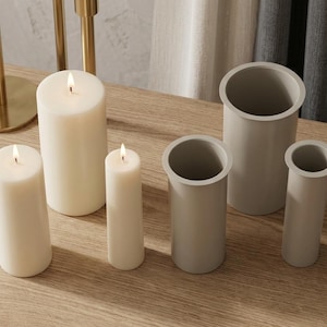 Puede incluir: Tres velas cilíndricas de cera blanca con llamas encendidas y tres recipientes cilíndricos grises de diferentes alturas con bordes anchos. Las velas y los recipientes están dispuestos en grupo y presentan un acabado liso y mate.