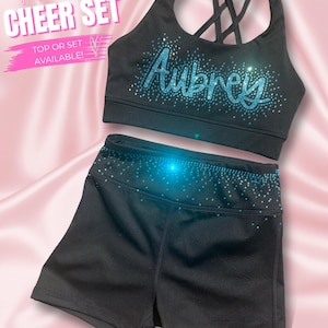 Peut inclure: Ensemble de cheerleading noir composé d'un soutien-gorge de sport et d'un short. Le soutien-gorge de sport a un dos croisé et le nom "Aubrey" en paillettes bleues. Le short et le soutien-gorge sont ornés de paillettes bleues. Le texte "Personalized Cheer Set" est visible.