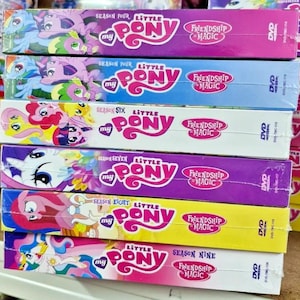 May include: A stack of DVD box sets for the animated series "My Little Pony: Friendship is Magic." Each box has a different colour scheme and features the show's logo and characters. Seasons four, six, seven, eight, and nine are visible.