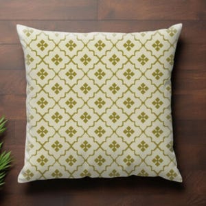 May include: Square decorative pillow with a cream-colored background and a repeating olive-green geometric pattern. The pattern consists of stylized four-leaf clover shapes. The pillow is placed on a dark brown wooden surface.