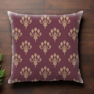 May include: A square decorative pillow with a burgundy background and a repeating gold floral pattern. The pillow is placed on a dark wood surface, with a green plant in the corner.