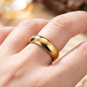 Puede incluir: Un anillo de banda en tono dorado pulido con una inscripción en escritura cursiva oscura y fluida grabada en toda la superficie exterior. El metal tiene un acabado liso y reflectante que capta la luz, resaltando las letras grabadas.