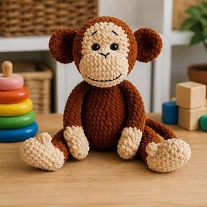 Op de afbeelding: Een handgemaakte gehaakte aap pop met bruin en beige garen. De aap heeft een lachend gezicht, grote oren en zit op een houten oppervlak. Andere speelgoed, waaronder een stapel kleurrijke ringen en houten blokken, zijn op de achtergrond.