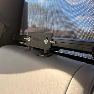 May include: A black car accessory is mounted on the back of a light gray car seat. The accessory has a rectangular shape with a black bar and is secured with bolts. A black wire extends from the accessory.