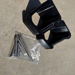 May include: Black plastic bracket with a rectangular opening and a clear plastic bag containing several metal screws and washers. The bracket appears to be 3D-printed.