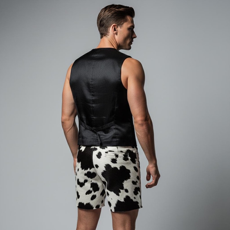 May include: A black vest and cowhide print shorts. The vest is sleeveless and has a satin finish. The shorts are white with black spots, resembling a cowhide pattern. The outfit is set against a gray backdrop.