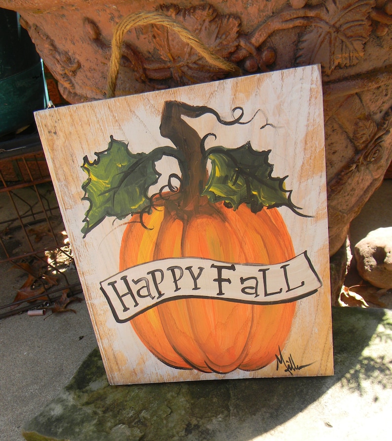 May include: A wooden sign with a painted pumpkin and the words "Happy Fall" in black script. The pumpkin is orange with green leaves and a brown stem.