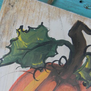 May include: A close-up of a painted wooden sign featuring a pumpkin with a green stem and leaves. The pumpkin is painted in shades of orange, yellow, and brown. The background is white with a wood grain texture.