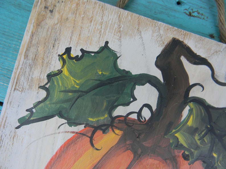 May include: A close-up of a painted wooden sign featuring a pumpkin with a green stem and leaves. The pumpkin is painted in shades of orange, yellow, and brown. The background is white with a wood grain texture.