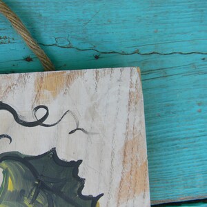 May include: A wooden sign with a white background and a painted design of a green leaf with black swirls. The sign is hanging on a turquoise blue wooden surface.