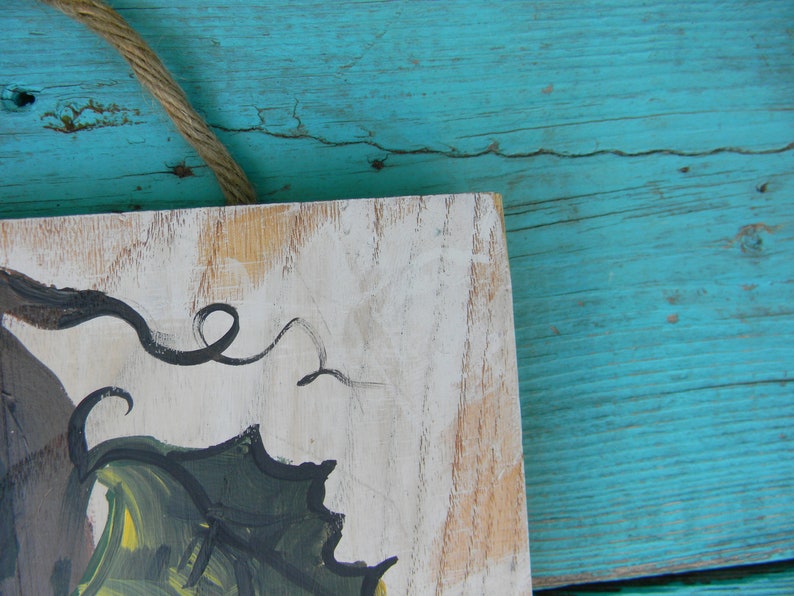May include: A wooden sign with a white background and a painted design of a green leaf with black swirls. The sign is hanging on a turquoise blue wooden surface.