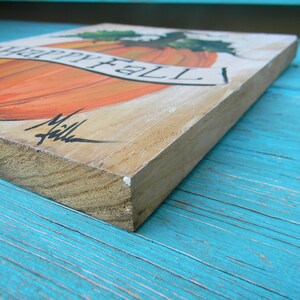 May include: A wooden sign with a painted orange pumpkin and the words "Happy Fall" in black lettering. The sign is on a blue wooden surface.
