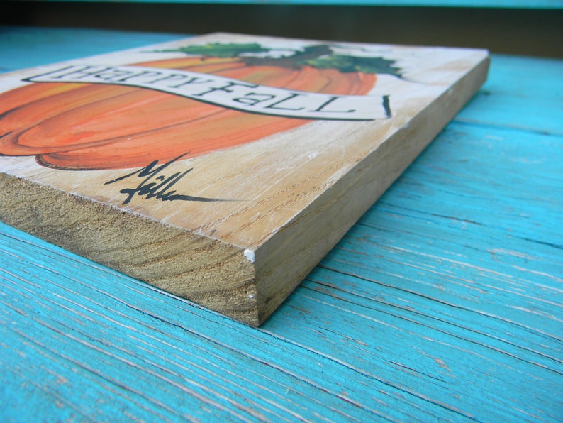 May include: A wooden sign with a painted orange pumpkin and the words "Happy Fall" in black lettering. The sign is on a blue wooden surface.