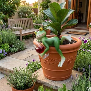 Puede incluir: Una maceta de terracota con una figura de rana verde sosteniendo una copa de vino tinto. La rana está posada en el borde de la maceta, que contiene una planta verde. La maceta tiene un diseño de vid.