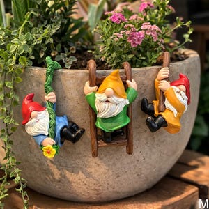 May include: A concrete planter pot filled with greenery and pink flowers. Three colorful garden gnomes are attached to the pot, one climbing a rope and two on ladders. The gnomes have white beards and wear red, yellow, and green outfits.