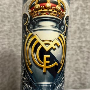 May include: A cylindrical tumbler featuring a gold crown and the Real Madrid CF emblem. The design includes a gold emblem with the letters "MCF" and a crown with red accents. The background has a dark, cloudy design.
