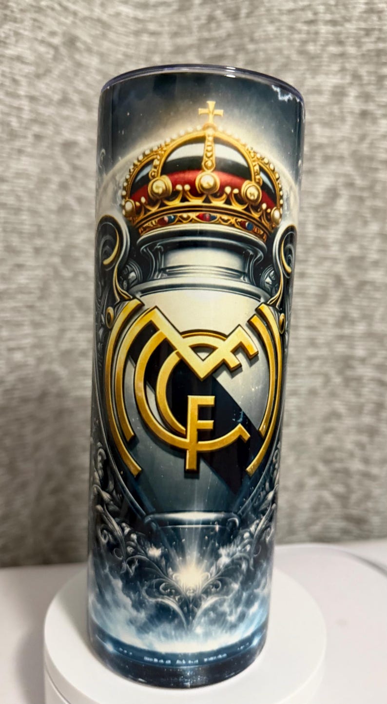 May include: A cylindrical tumbler featuring a gold crown and the Real Madrid CF emblem. The design includes a gold emblem with the letters "MCF" and a crown with red accents. The background has a dark, cloudy design.