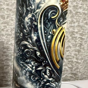 May include: A cylindrical tumbler with a detailed design. The artwork features a crown, shield, and ornate embellishments in gold and silver tones against a stormy sky background. The overall theme is regal and decorative.