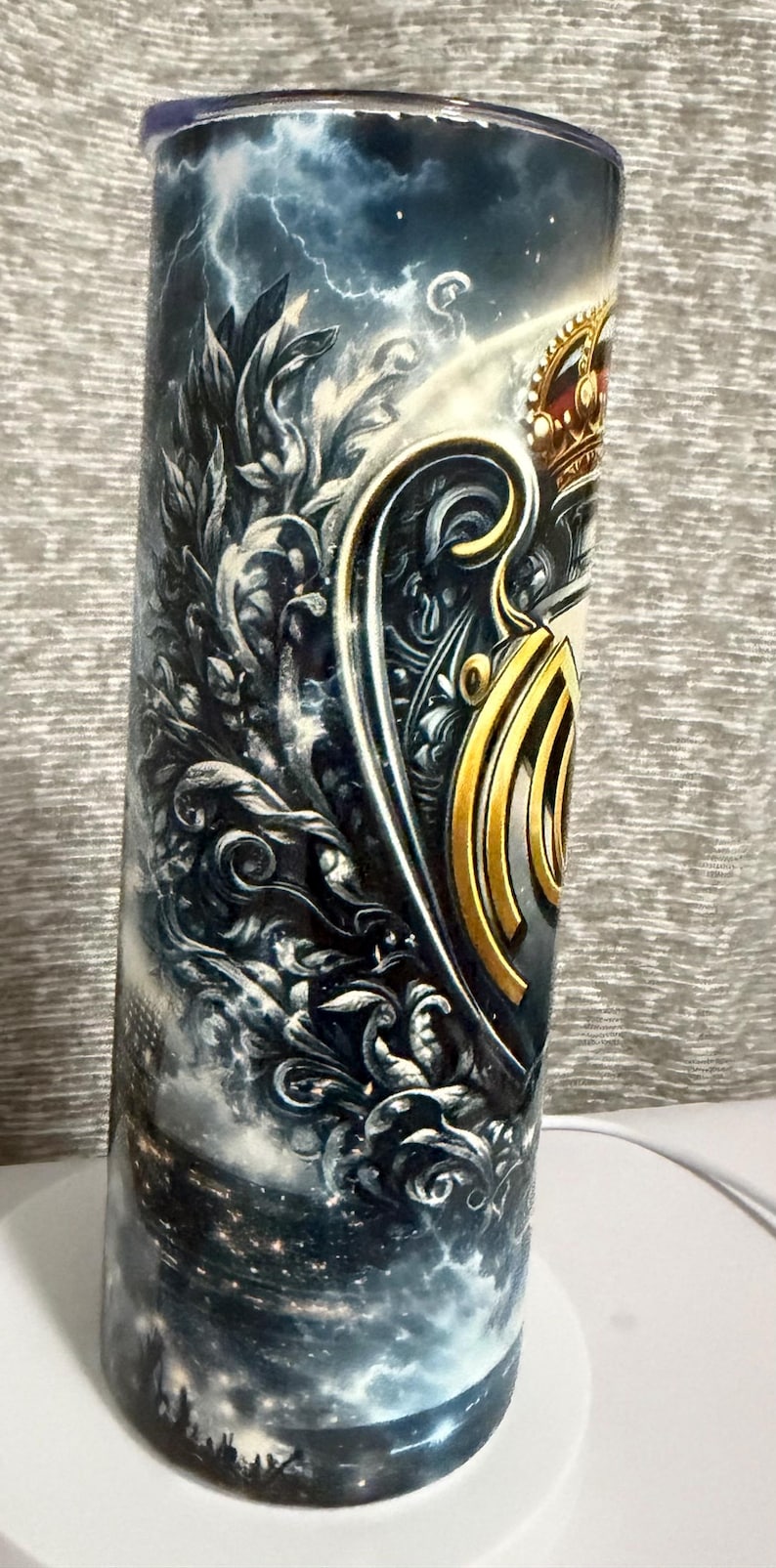 May include: A cylindrical tumbler with a detailed design. The artwork features a crown, shield, and ornate embellishments in gold and silver tones against a stormy sky background. The overall theme is regal and decorative.
