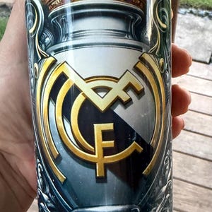 May include: A cylindrical tumbler featuring the Real Madrid CF logo in gold and white, set against a dark blue background. The design includes a crown and ornate detailing, creating a regal aesthetic. The tumbler is held by a hand.