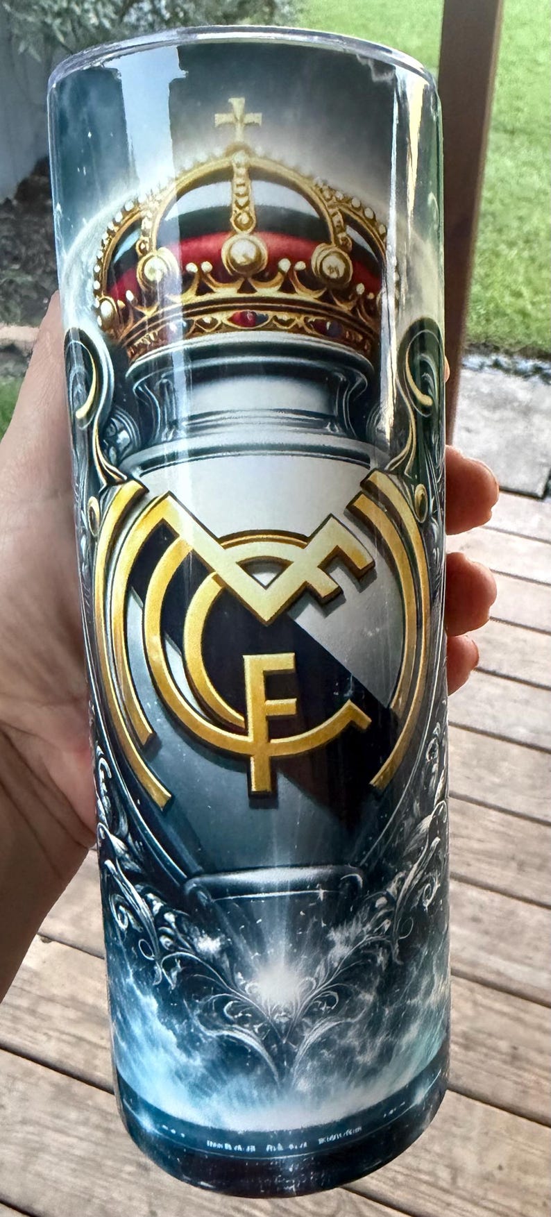May include: A cylindrical tumbler featuring the Real Madrid CF logo in gold and white, set against a dark blue background. The design includes a crown and ornate detailing, creating a regal aesthetic. The tumbler is held by a hand.