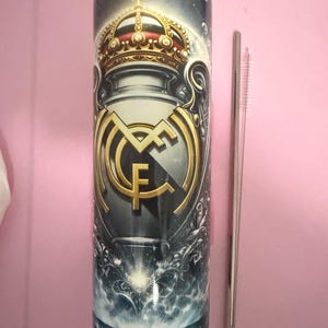 May include: A cylindrical tumbler featuring the Real Madrid crest in gold and white against a dark background. The design includes a crown and ornate details. A silver straw and cleaning brush are next to the tumbler.