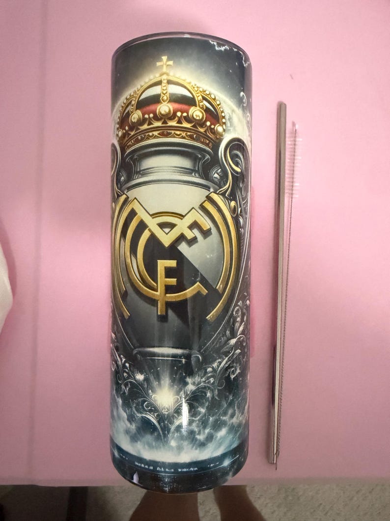 May include: A cylindrical tumbler featuring the Real Madrid crest in gold and white against a dark background. The design includes a crown and ornate details. A silver straw and cleaning brush are next to the tumbler.