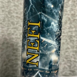 May include: A stainless steel tumbler with a wraparound design featuring a dramatic thunderstorm scene with lightning bolts and a stadium crowd. The word "NEH" is printed vertically in gold letters.