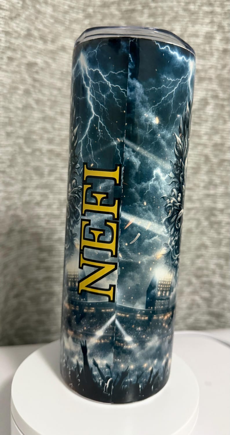 May include: A stainless steel tumbler with a wraparound design featuring a dramatic thunderstorm scene with lightning bolts and a stadium crowd. The word "NEH" is printed vertically in gold letters.