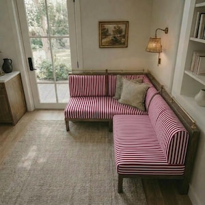 Könnte beinhalten: Ein L-förmiges Sofa mit Holzrahmen und Kissen mit vertikalen rot-weißen Streifen. Das Sofa befindet sich in einer Ecke eines Raumes mit einem neutralen Teppich und einem Fenster. Ein kleines Gemälde hängt an der Wand.